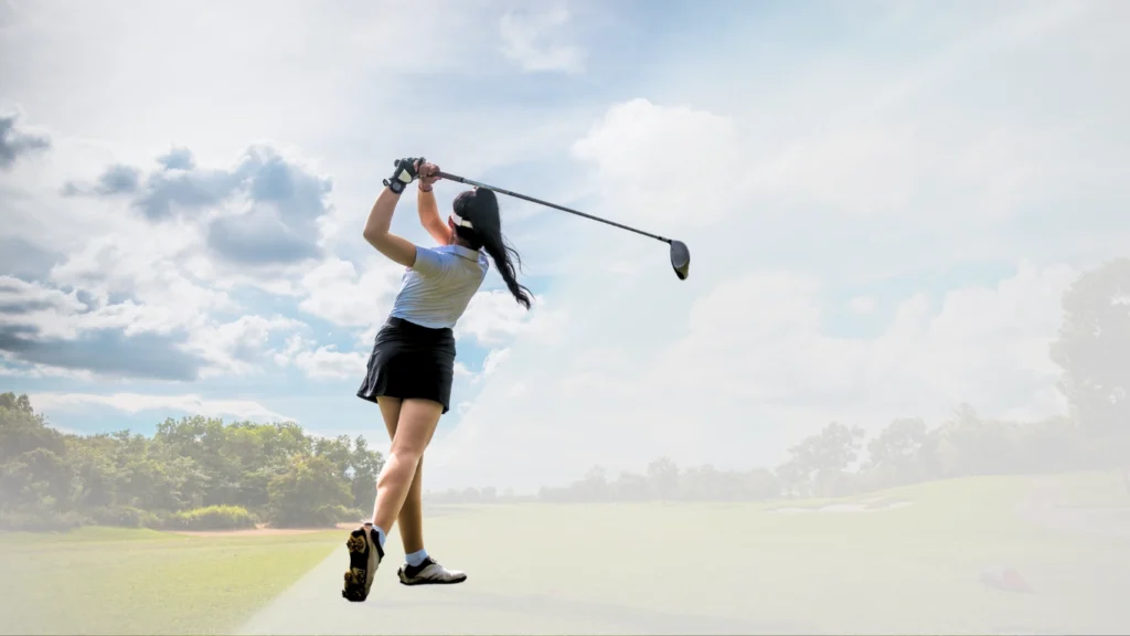 Golfista de costas no ápice de um swing em um campo de golfe verdejante, sob um céu azul com nuvens. Ela veste camisa de gola polo branca e saia preta, e o taco de golfe está estendido no final do movimento. A imagem possui um filtro suave e uma sobreposição branca na parte inferior.