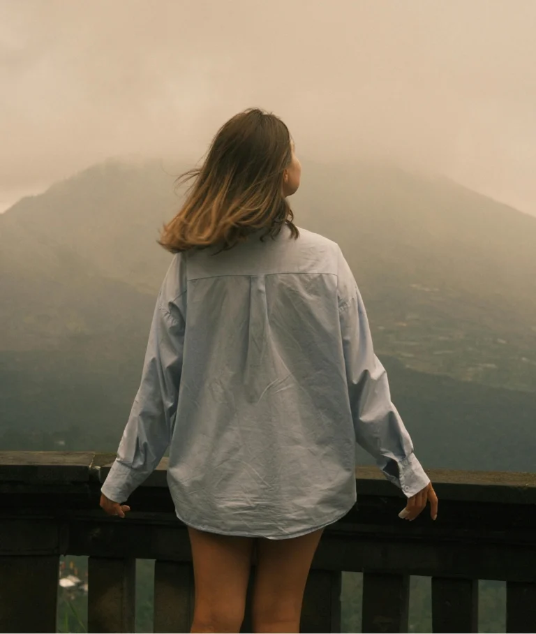 Mulher de costas em mirante de pedra observando vasta paisagem montanhosa envolta em névoa suave. Ela veste camisa de botão azul claro oversized e tem cabelos castanhos. O fundo é nebuloso e inspirador.