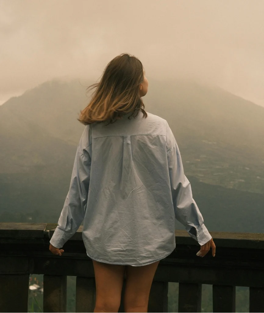 Mulher de costas em mirante de pedra observando vasta paisagem montanhosa envolta em névoa suave. Ela veste camisa de botão azul claro oversized e tem cabelos castanhos. O fundo é nebuloso e inspirador.