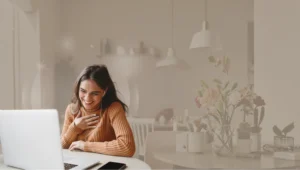 Mulher sorridente de blusa de tricô cor mostarda sentada à mesa, interagindo em uma videochamada no notebook. O ambiente ao fundo é uma sala clara com flores em um vaso e luminárias pendentes, tudo sob um filtro suave em tons de bege.