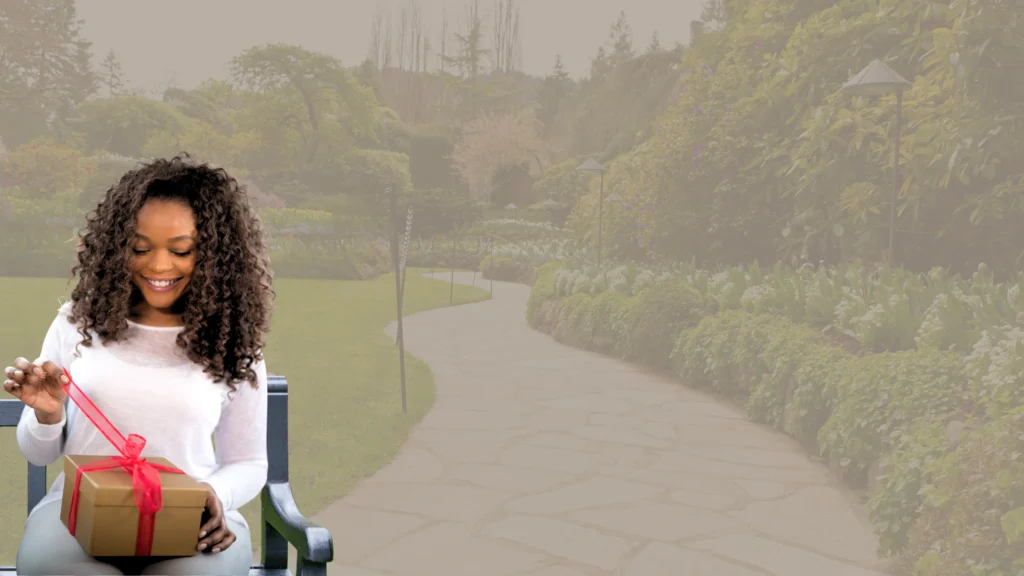 Mulher negra de cabelos cacheados, sorrindo e sentada em um banco de madeira, abre uma caixa de presente dourada com um laço vermelho. Ao fundo, um jardim com um caminho de pedra e muitas plantas verdes, com uma luz suave e desfocada.