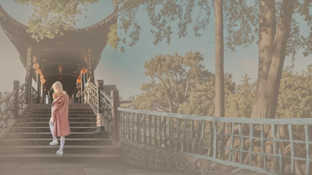 Mulher loira de costas, vestindo sobretudo rosa e tênis brancos, subindo os degraus de uma ponte de madeira em estilo oriental. O ambiente é arborizado e banhado por uma luz solar suave e amarelada.