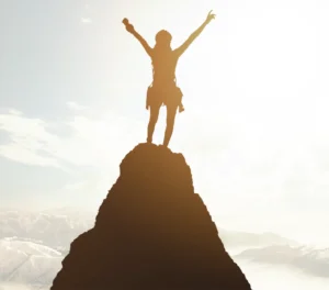 Silhueta de uma mulher no topo de uma montanha rochosa com os braços erguidos em sinal de vitória. Ela usa equipamentos de escalada. O sol brilha intensamente ao fundo, criando uma luz dourada difusa sobre um céu claro com nuvens sutis.