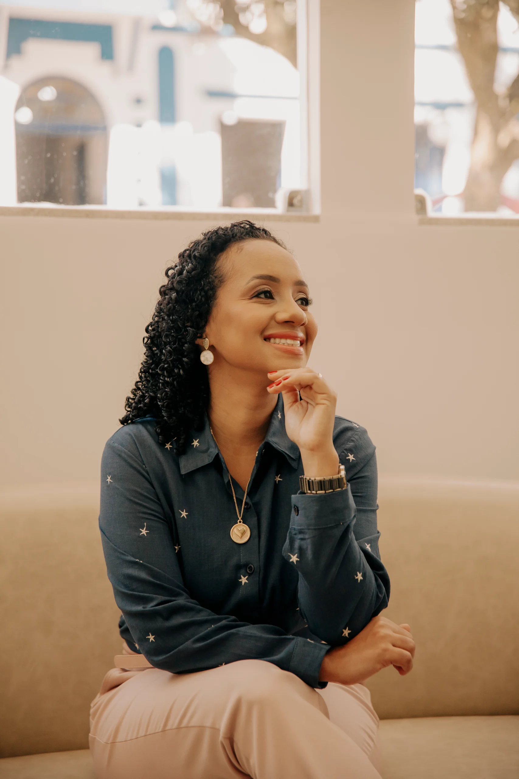 Retrato da psicóloga Lívia Dias, negra com cabelos cacheados e sorriso genuíno, sentada em um sofá bege em consultório bem iluminado. Ela veste blusa azul-marinho com estrelas sutis, calça clara e olha levemente para o lado com a mão no queixo. O fundo tem janelas amplas com luz natural e vista urbana desfocada.